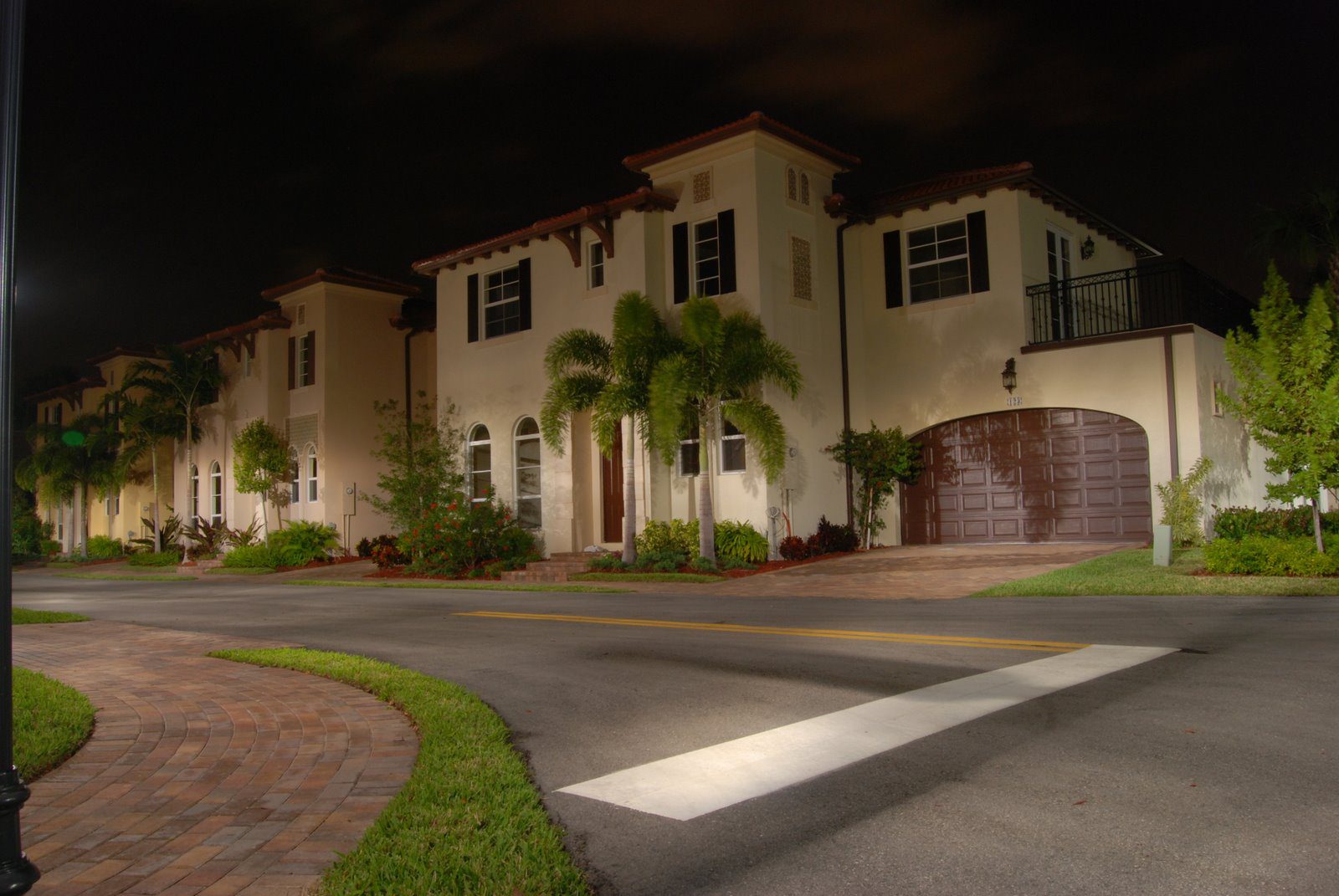 A nighttime view of a modern house with palm trees and well-lit driveway.