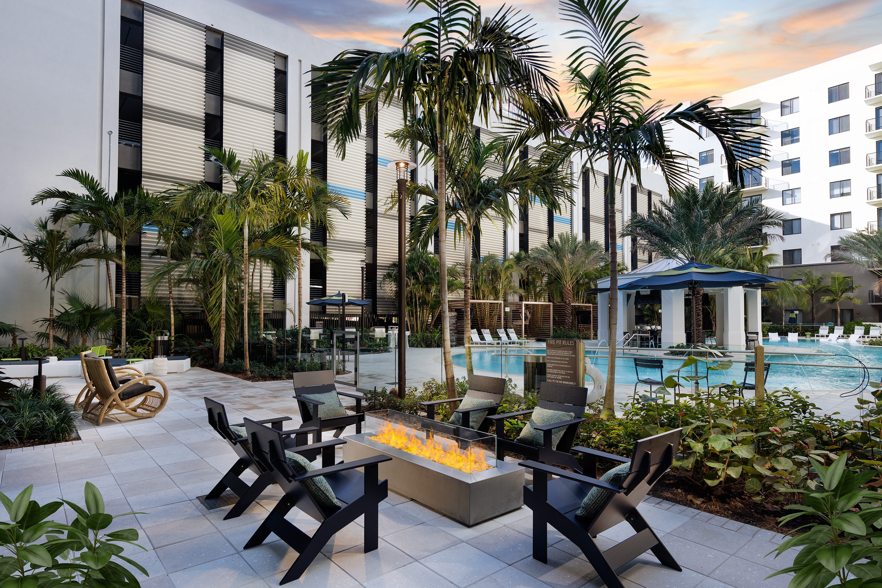 Outdoor seating area with firepit near a pool and palm trees at sunset.
