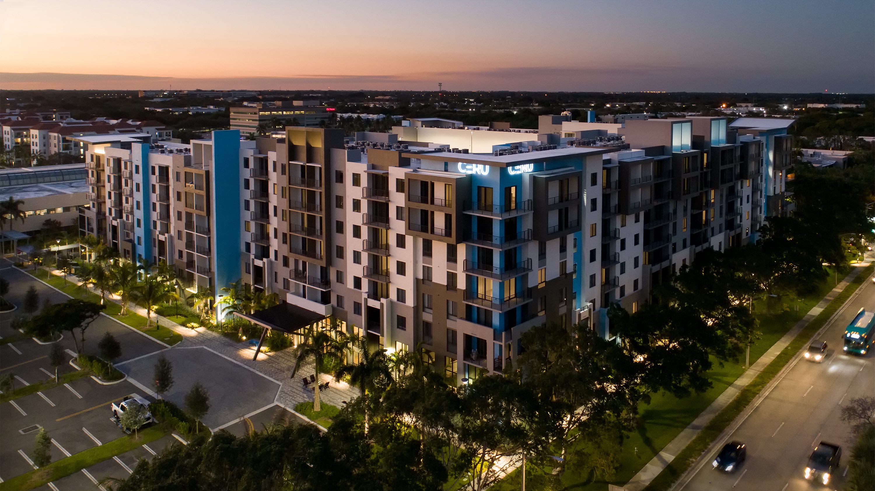 Modern multi-story apartment building at dusk with illuminated windows and surrounding greenery.