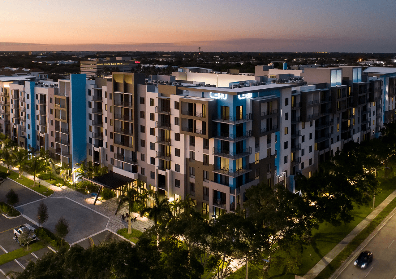 Modern apartment complex at dusk with illuminated streets and greenery.