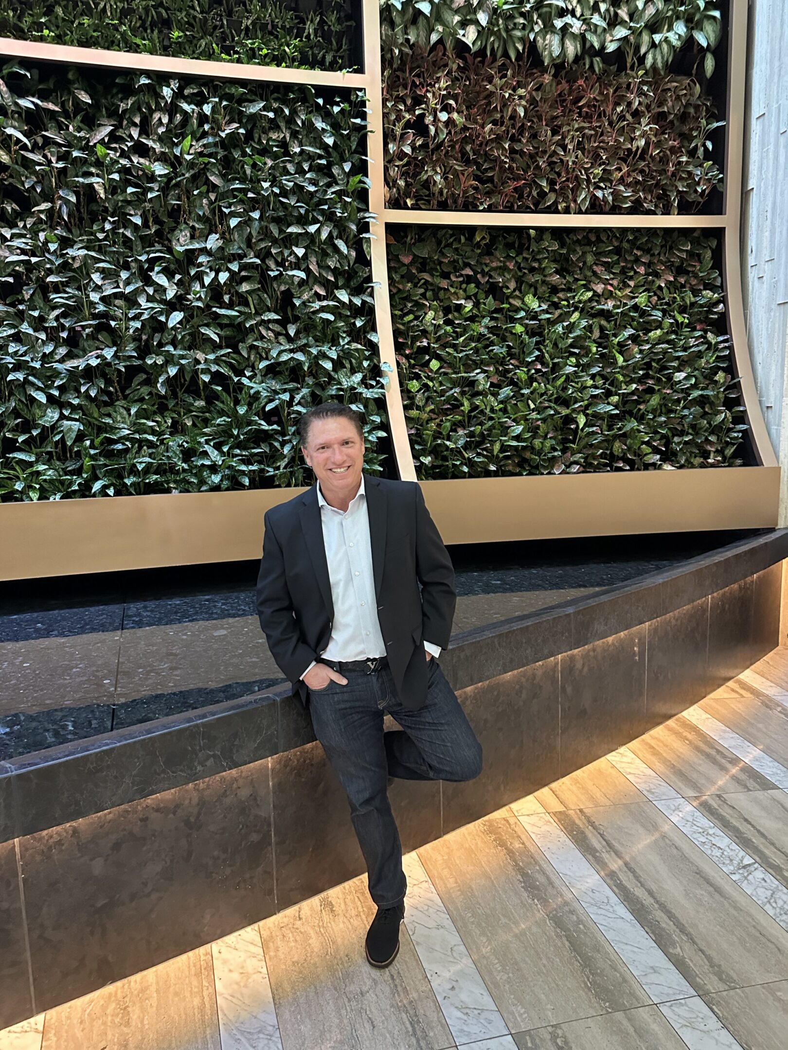 Man in black suit posing indoors near a green wall.